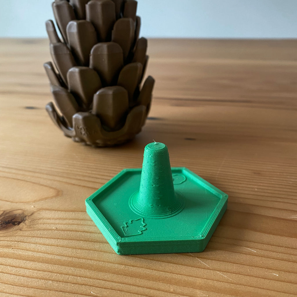 The Stopple Stand is sitting on a wooden table with the Loblolly Pinecone Puzzle Toy behind it. The Stopple Stand is green.