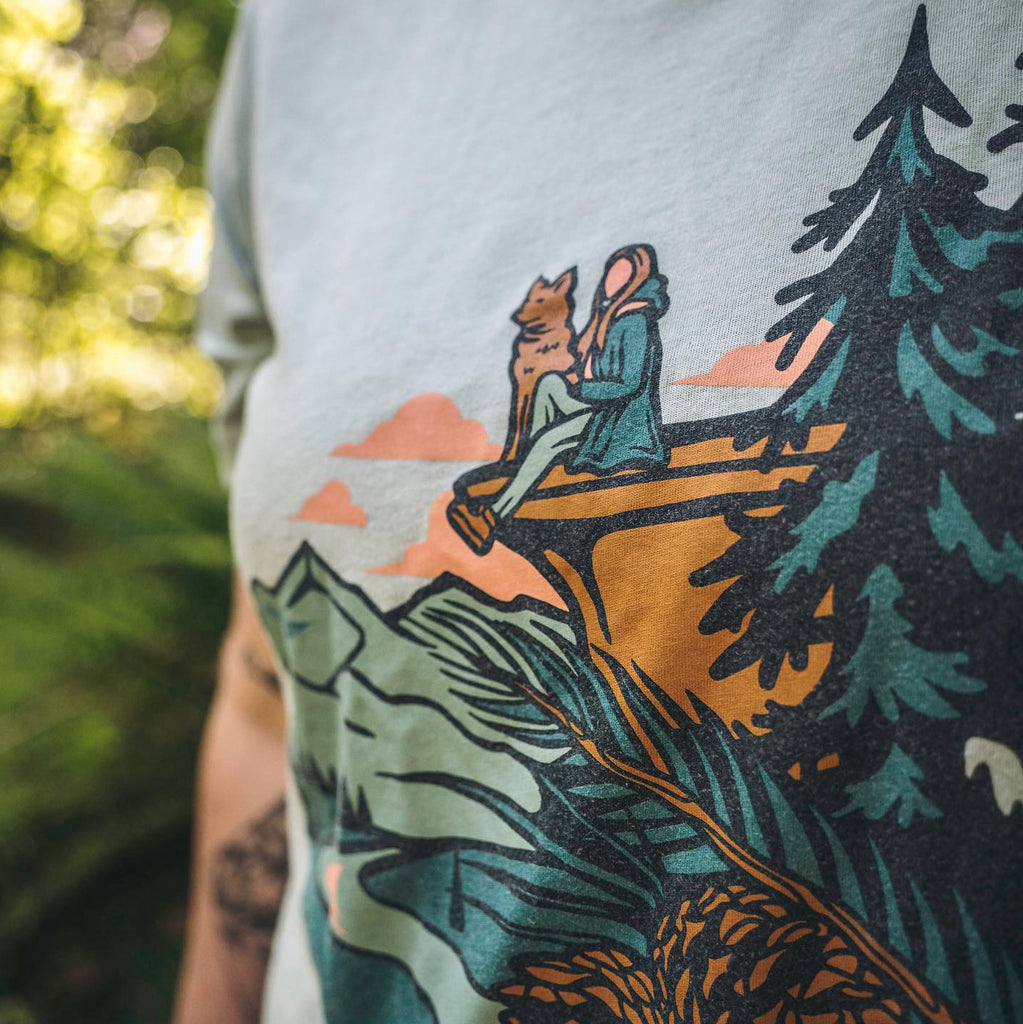 A woman wearing the Dog at Dusk Shirt by Loblola. The shirt shows a woman sitting on a cliff with her dog. There are pine trees, mountains, and pink clouds around.