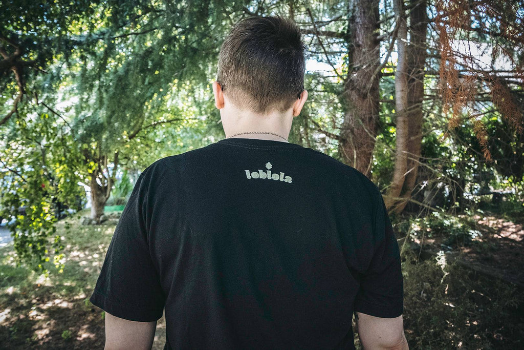 Woman standing in the woods facing away. The back of her black shirt shows the Loblola logo in white.