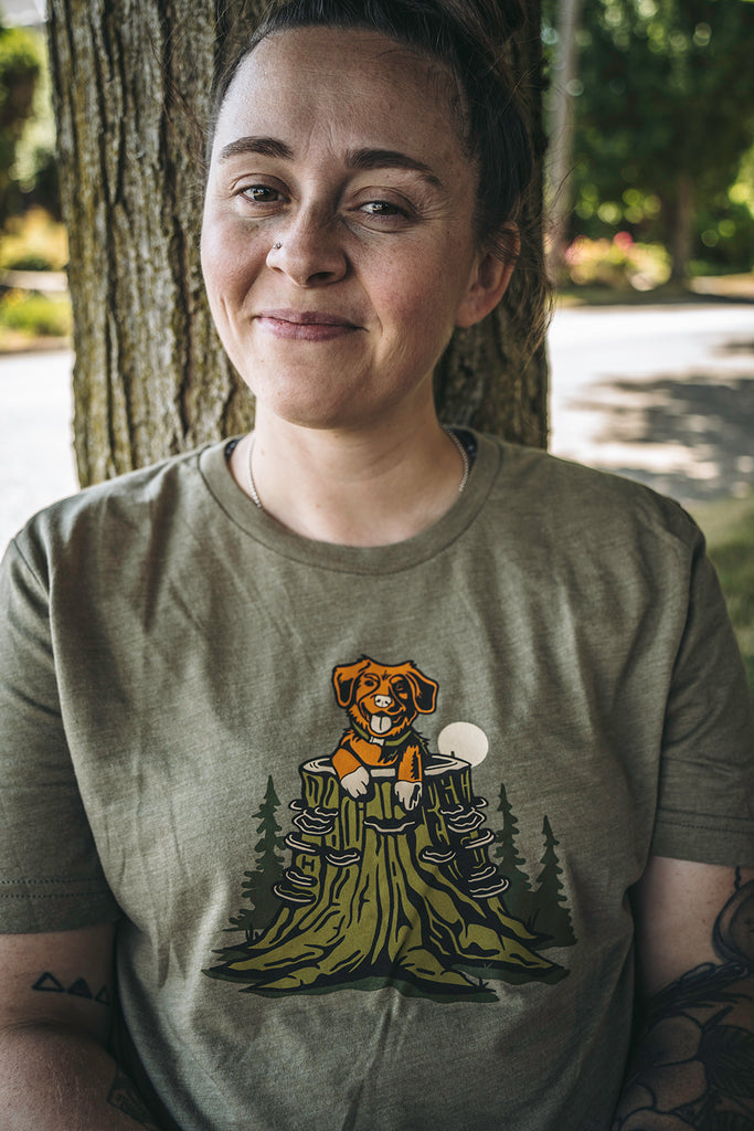 Woman leaning against a tree trunk wearing the Pup in a Stump shirt from Loblola. She is smiling. The shirt shows an orange dog poking up out of a green tree trunk with turkey tail mushrooms.
