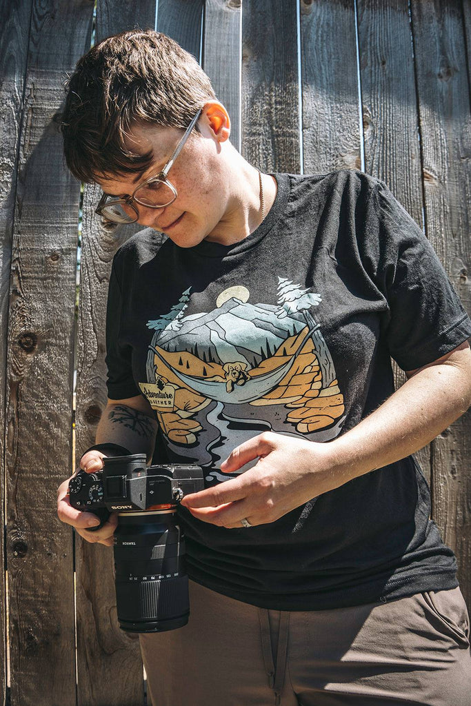 Woman leaning against a fence and looking at her camera. She is wearing the black Adventure Together shirt from Loblola.
