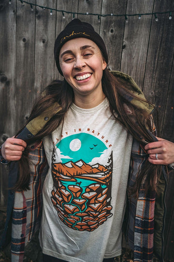 A woman is pulling open her jacket to show off the Play in Nature Tee Shirt by Loblola. The shirt is an oat color with a design that shows a dog in a river with mountains in the background.