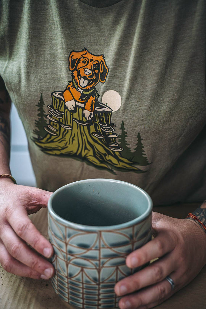 Close up of a woman wearing the Pup in a Stump shirt from Loblola. She is smiling. The shirt shows an orange dog poking up out of a green tree trunk with turkey tail mushrooms