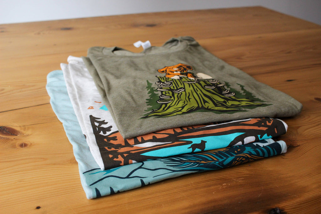 Stack of Loblola Nature Dog graphic tees on a wooden table. The shirts are green, tan, and blue.