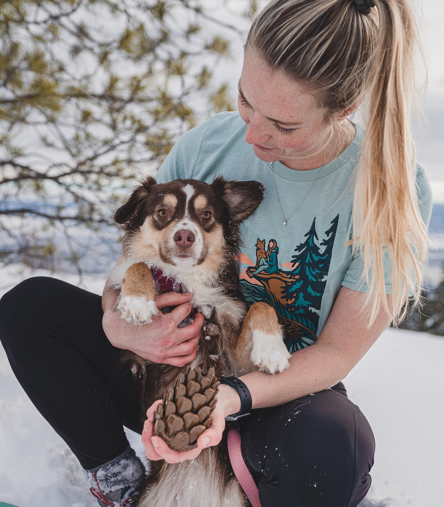 Woman and dog outside in the snow. The woman is wearing the Loblola Dog at Dusk shirt and holding the Loblolly Pinecone Dog Toy.
