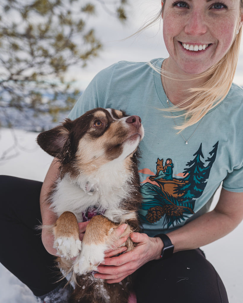 Woman outside wearing Loblola Dog at Dusk shirt which features an illustrated woman with her dog sitting on a cliff amongst the pine trees.
