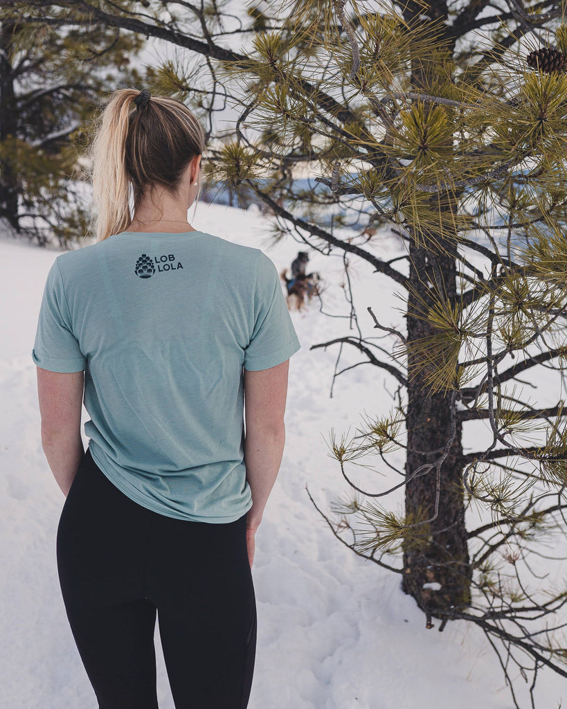 Woman outside in the snow wearing the Loblola Dog at Dusk tshirt which features an illustrated woman with her dog sitting on a cliff. The back shows the Loblola logo.
